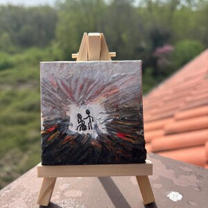 Può includere: Piccolo dipinto quadrato su un cavalletto di legno. L'opera raffigura due figure stilizzate in una scena oscura e astratta con accenti rossi e bianchi. Lo sfondo è una miscela di grigio e bianco.