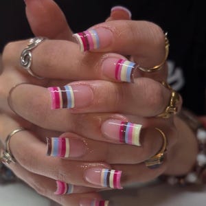Puede incluir: Uñas artificiales de forma cuadrada con estilo de manicura francesa y rayas horizontales en rosa, marrón, azul claro y crema en las puntas. Los dedos lucen varios anillos de oro y plata, presentando un acabado brillante y pulido.
