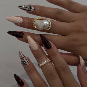 May include: Stiletto-shaped nails featuring a mix of glossy dark brown polish, beige cable-knit texture, and a beige plaid pattern. One finger wears a large, irregular-shaped white pearl ring in a gold setting, while another features a gold band with clear stones.