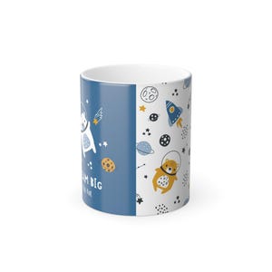 May include: White ceramic mug with a whimsical space-themed design. One side features a blue background with a bear astronaut and the text "Dream Big". The other side has a pattern of planets, stars, and rockets. The mug is perfect for coffee or tea.