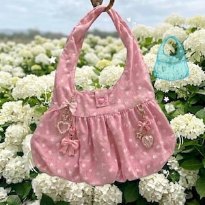 May include: Pink polka-dot handbag with a curved handle, hanging from a bronze hook. The bag features a ruffled bottom, a heart-shaped button closure, and decorative charms with bows and hearts. A light blue handbag sketch is in the background.