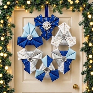 Op de afbeelding: Een cirkelvormige krans bestaande uit vijf gevouwen stoffen ornamenten in blauwe, zilveren en witte tinten. Elk ornament heeft een centrale knoop, sommige versierd met witte kunstparels. De ornamenten zijn in een ring gerangschikt en opgehangen.