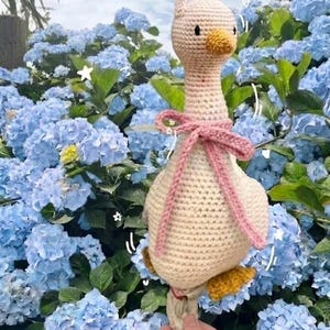 May include: A handmade crochet goose toy with a cream-colored body, a yellow beak and feet, and a pink bow. The goose is suspended against a backdrop of blue hydrangea flowers. The toy is designed with a whimsical and charming aesthetic.