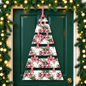Puede incluir: Una decoración de pared colgante con forma de árbol de Navidad, compuesta por cinco listones de madera blancos horizontales con un patrón de ramas de pino verdes, bayas rojas y lazos rosas, unidos por cintas rosas y un anillo dorado.