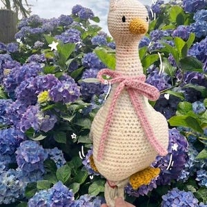 May include: Crocheted goose doll with a cream-colored body, yellow feet, and a pink bow around its neck. The goose has a yellow beak and is hanging from a brass hook. The background features vibrant purple hydrangea flowers.