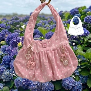 Könnte beinhalten: Eine rosa Handtasche mit weißen Punkten, mit einem geschwungenen Griff, verziert mit Schleifen- und herzförmigen Anhängern. Die Tasche hat einen Knopfverschluss mit Herzdetail und einen gerafften Boden. Im Hintergrund lila Hortensien.