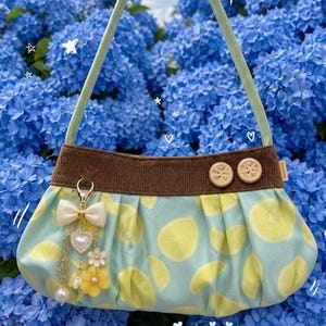 Könnte beinhalten: Hellblaue Handtasche mit Zitronenmuster, braunem Besatz und grünem Griff. Die Tasche hat Holzknöpfe und einen Anhänger mit Schleife, Herz und Blumen. Der Hintergrund ist ein Feld blauer Blumen.