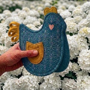 May include: A teal chicken-shaped pot holder with a gold comb, wing, and pocket. The pot holder has a patterned design and a small pink heart. The pot holder is made of felt.
