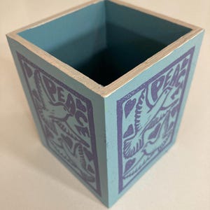 May include: A teal-painted square wooden box with silver trim. The sides display a repeating pattern of a dove and the word "PEACE" in purple. The inside of the box is teal.
