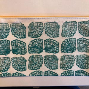 May include: A rectangular, off-white canvas pencil case with a repeating pattern of teal seashells. The case has a yellow zip and a gold-coloured zip pull. The design is a coastal or nautical theme.
