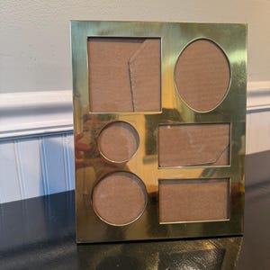 May include: A rectangular gold-toned metal multi-photo frame featuring a polished finish. The frame contains five distinct openings for photographs: one large vertical rectangle, one oval, one small circle, and two horizontal rectangles.