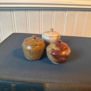 May include: Three small, lidded jars in shades of brown, tan, and red-brown sit on a blue book. The jars have a rounded shape with a small knob on top of the lid. The jars appear to be made of stone or a similar material.
