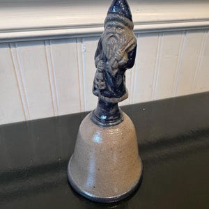 May include: A ceramic bell featuring a sculpted figure of a bearded person wearing a pointed hat as the handle. The figure is finished in a dark blue glaze, while the bell body has a speckled tan finish with a dark blue rim at the base.