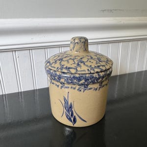 Può includere: Un barattolo cilindrico in gres con coperchio abbinato, caratterizzato da una base color sabbia con dettagli blu spugnati sul coperchio e sul bordo. Un disegno di spiga di grano dipinto a mano in blu è centrato sulla parte anteriore.
