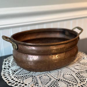 May include: An oval-shaped copper basin featuring a hammered texture and two weathered brass handles attached to the sides. The vessel has a wide, open top and a rounded base, displaying a natural patina with variations in the metallic finish.