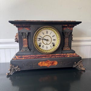 May include: A black mantel clock with faux marble accents in orange and brown. The clock features a circular face with Roman numerals, a gold-toned bezel, and two black hands. It is supported by four ornate metal feet and has columns.