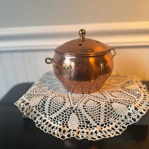 May include: A polished copper pot with a rounded body, two small side handles, and a matching domed lid featuring a central brass knob and small ventilation holes. The pot has a smooth, reflective surface and a warm metallic finish throughout.