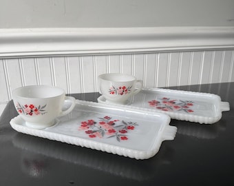 May include: Two white milk glass snack sets, each consisting of a rectangular tray with a scalloped edge and a matching teacup. Both pieces feature a delicate floral pattern in shades of red, pink, and grey, with a circular indentation on the tray.
