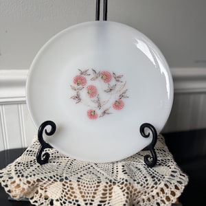 May include: A white plate with a floral design featuring pink flowers and brown leaves. The plate is displayed on a black metal stand with a decorative scroll design, resting on a cream-colored lace doily. A vintage-style decorative piece.