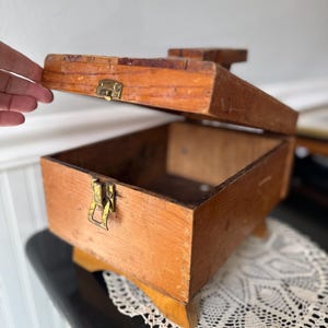 Puede incluir: Una pequeña caja de madera abierta con tapa con bisagras. La caja es de madera marrón claro y tiene un cierre de latón. La caja está sobre una pequeña base de madera. Una mano está abriendo la tapa.