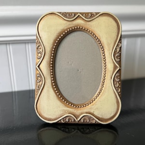 May include: A rectangular picture frame with an oval center opening. The frame features a cream-colored face with distressed gold-toned edges and intricate embossed scrollwork details. A beaded border surrounds the central oval aperture.