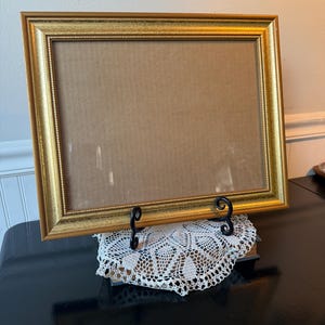 May include: A rectangular gold picture frame with a decorative border. The frame is propped up on a black metal stand, resting on a white lace doily. The frame is empty, with a cardboard backing.