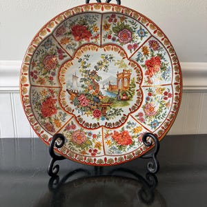Puede incluir: Bandeja decorativa redonda con un diseño floral y una escena de paisaje central. La bandeja tiene un borde rojo y está sostenida por un soporte de metal negro. El patrón floral incluye flores rojas, rosas y amarillas.