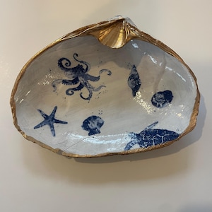 May include: A decorative seashell dish with a gold-painted rim. The interior is white with blue illustrations of an octopus, starfish, seashells, and a turtle. A unique coastal-themed accent piece.