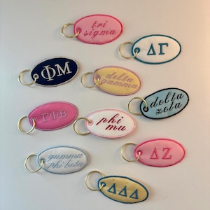 May include: A collection of ten oval-shaped embroidered keychains featuring various Greek letters and sorority names including tri sigma, delta gamma, delta zeta, phi mu, gamma phi beta, and others, each attached to a gold-toned metal split ring.
