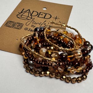 May include: A stack of multi-strand beaded bracelets featuring a variety of brown, amber, and metallic gold beads in different shapes and sizes. The bracelets are displayed on a brown cardboard tag with the text JADED Jewelz printed in black.
