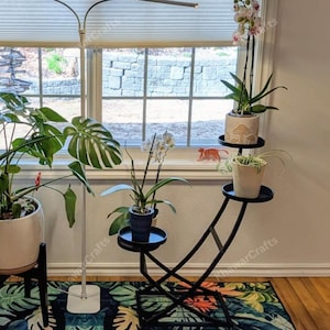 May include: A black metal plant stand with three circular shelves, holding potted plants with green foliage and white flowers. The stand has a curved design and is positioned near a window with natural light.