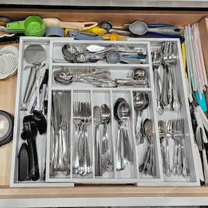 May include: A white drawer organizer filled with silverware, including forks, knives, and spoons. Various kitchen utensils, such as measuring spoons and a pizza cutter, are also visible. The drawer is open, revealing the organized contents.