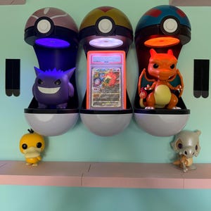 Könnte beinhalten: Drei dekorative Regale in Form von Poké-Bällen, die jeweils eine andere Pokémon-Figur oder -Karte enthalten. Die Regale sind an einer hellblauen Wand befestigt, darunter ein rosa Regal mit zusätzlichen Figuren. Die Regale sind weiß und haben ein schwarz-weißes Design.