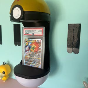 May include: A wall-mounted display stand shaped like a gold and white Poke Ball holding a graded Charizard VSTAR trading card in a clear plastic PSA slab. The card features a red dragon illustration and the text 2022 POKEMON SWSH BSP #262.