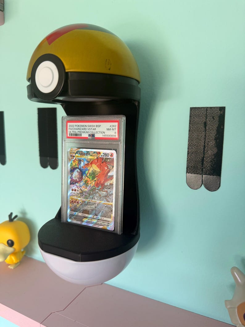 May include: A wall-mounted display stand shaped like a gold and white Poke Ball holding a graded Charizard VSTAR trading card in a clear plastic PSA slab. The card features a red dragon illustration and the text 2022 POKEMON SWSH BSP #262.