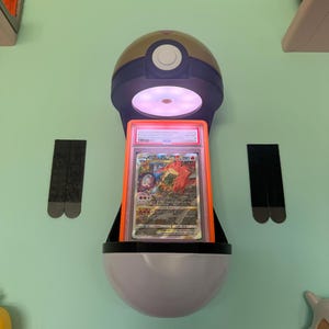 Pode incluir: Um expositor de parede em forma de Poké Bola, contendo um cartão colecionável classificado. O expositor é principalmente branco, preto e dourado, com uma luz que ilumina o cartão. O cartão apresenta obras de arte coloridas.