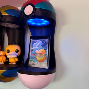 May include: A wall-mounted display shelf in the shape of a Poké Ball, with a blue LED light and a Pokémon trading card. The shelf is black, blue, red, and white, with a pink base. Other similar shelves are visible.