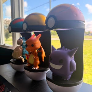 Op de afbeelding: Een verzameling Pok&eacute;mon-beeldjes, waaronder Charizard en Gengar, op zwarte standaards met witte bases. Elke figuur staat onder een Pok&eacute; Ball-vormige lamp. De achtergrond toont een raam.