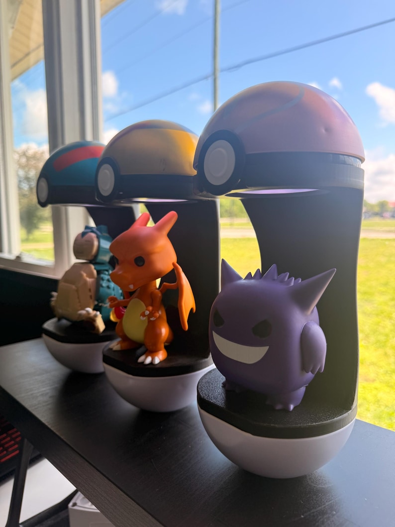 Op de afbeelding: Een verzameling Pok&eacute;mon-beeldjes, waaronder Charizard en Gengar, op zwarte standaards met witte bases. Elke figuur staat onder een Pok&eacute; Ball-vormige lamp. De achtergrond toont een raam.