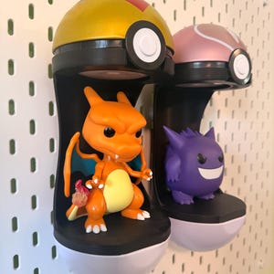Op de afbeelding: Twee displayrekken met Pok&eacute;mon-thema. E&eacute;n plank bevat een oranje Charizard-figuur en de andere een paarse Gengar-figuur. De planken zijn zwart en wit, met een geel en roze Pokeball-ontwerp erbovenop.