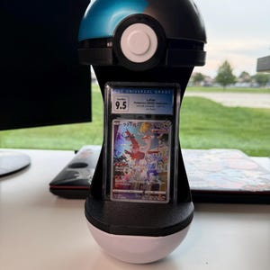 May include: A black and blue spherical display stand shaped like a Poke Ball, holding a vertically oriented, rectangular CGC graded trading card. The card features a Latias illustration with Japanese text and a Gem Mint 9.5 grade label at the top.