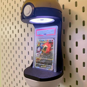 May include: A wall-mounted display stand shaped like a gold and white Pok&eacute; Ball featuring an integrated LED light that illuminates a graded Charizard VSTAR trading card encased in a clear plastic PSA slab with a white label and black text.
