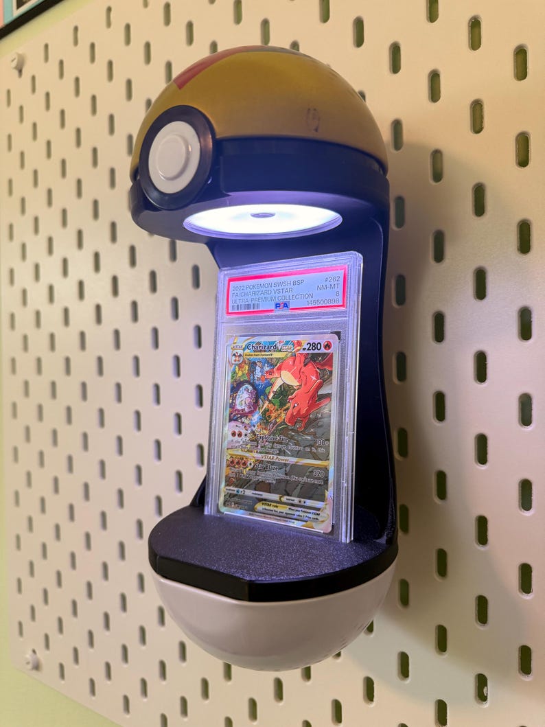 May include: A wall-mounted display stand shaped like a gold and white Pok&eacute; Ball featuring an integrated LED light that illuminates a graded Charizard VSTAR trading card encased in a clear plastic PSA slab with a white label and black text.