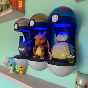 Op de afbeelding: Drie aan de muur gemonteerde Pok&eacute; Ball-vormige displaykasten met blauwe LED-verlichting, elk met een ander Pok&eacute;mon-figuur: Gengar, Charizard en Snorlax. Extra Pok&eacute;mon-figuren worden op een plank hieronder weergegeven.