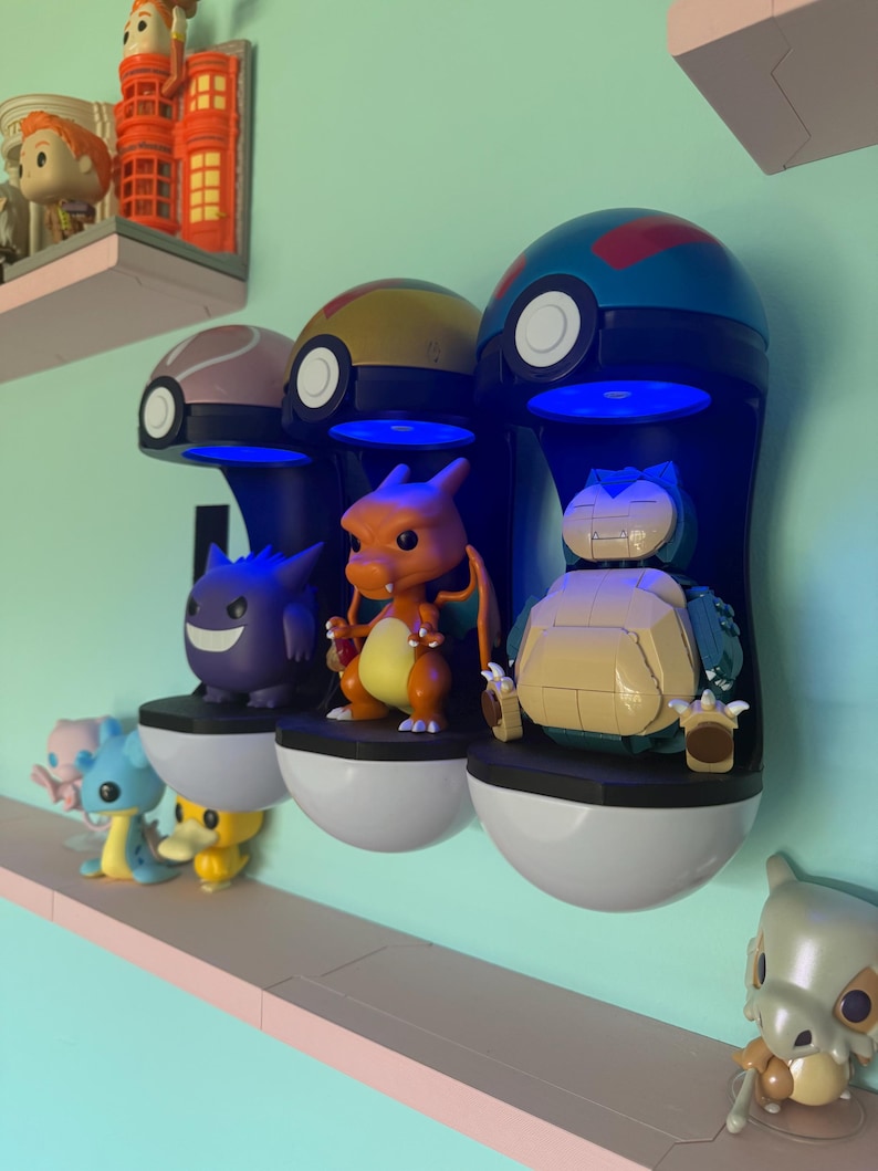 Op de afbeelding: Drie aan de muur gemonteerde Pok&eacute; Ball-vormige displaykasten met blauwe LED-verlichting, elk met een ander Pok&eacute;mon-figuur: Gengar, Charizard en Snorlax. Extra Pok&eacute;mon-figuren worden op een plank hieronder weergegeven.