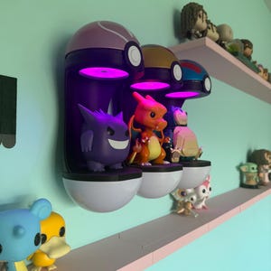 Op de afbeelding: Een display van Pok&eacute;mon-thema verzamelobjecten. Drie aan de muur gemonteerde Pok&eacute; Ball-vormige planken met paarse, gouden en blauwe toppen houden beeldjes vast. Een paarse Gengar, oranje Charizard en andere figuren worden getoond. Extra planken bevatten meer beeldjes.