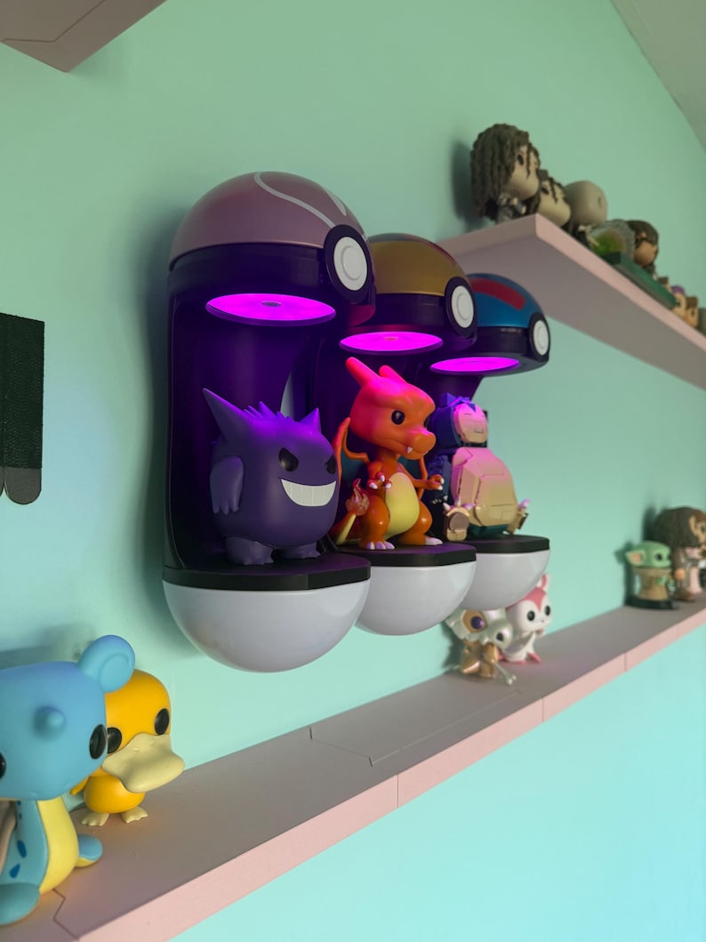 Op de afbeelding: Een display van Pok&eacute;mon-thema verzamelobjecten. Drie aan de muur gemonteerde Pok&eacute; Ball-vormige planken met paarse, gouden en blauwe toppen houden beeldjes vast. Een paarse Gengar, oranje Charizard en andere figuren worden getoond. Extra planken bevatten meer beeldjes.