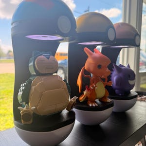 Op de afbeelding: Drie Pok&eacute;mon-displaystandaards. Elke standaard heeft een zwarte basis met een witte onderste helft, bekroond met een gekleurde koepel. De standaards bevatten figuren van Snorlax, Charizard en Gengar. De standaards worden op een zwarte plank weergegeven.