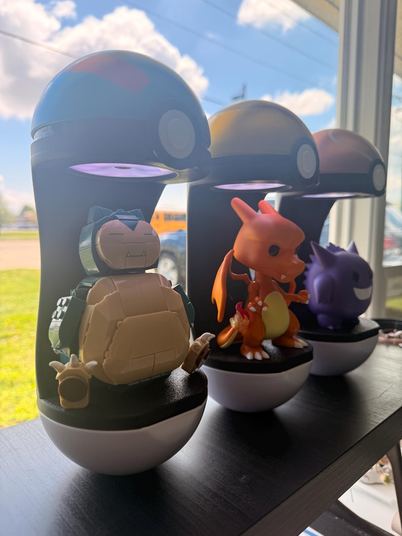 Op de afbeelding: Drie Pok&eacute;mon-displaystandaards. Elke standaard heeft een zwarte basis met een witte onderste helft, bekroond met een gekleurde koepel. De standaards bevatten figuren van Snorlax, Charizard en Gengar. De standaards worden op een zwarte plank weergegeven.