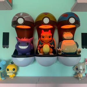 Op de afbeelding: Drie Pok&eacute;mon-thema displayplanken, elk met een ander kleurenschema, bevatten karakterbeeldjes. De planken zijn gevormd als Pok&eacute; Balls, met een licht dat van bovenaf schijnt. De beeldjes zijn onder andere Gengar, Charizard en Snorlax.